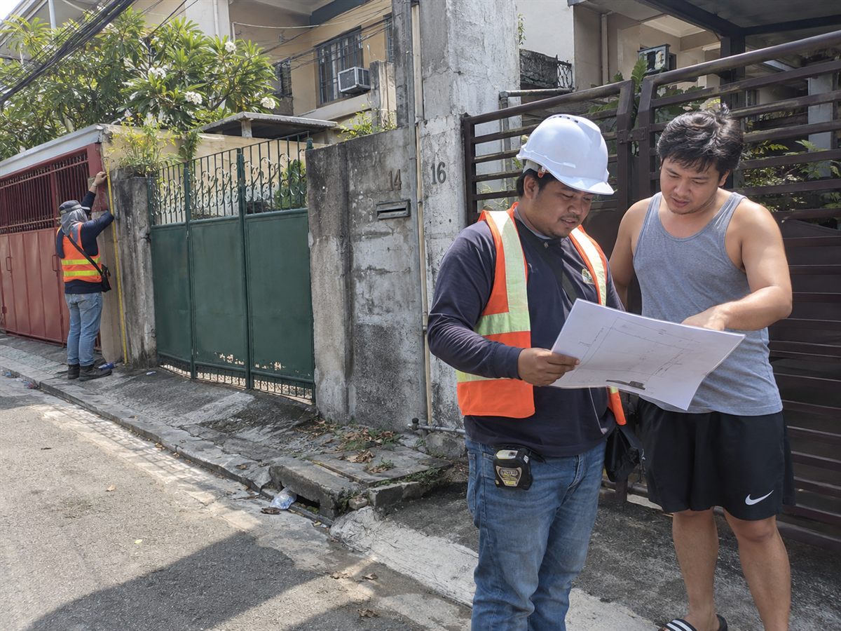 Surveyor in an orange safety vest and hard hat reviewing a survey plan with a homeowner outside a gate, while a crew member measures along the wall behind them