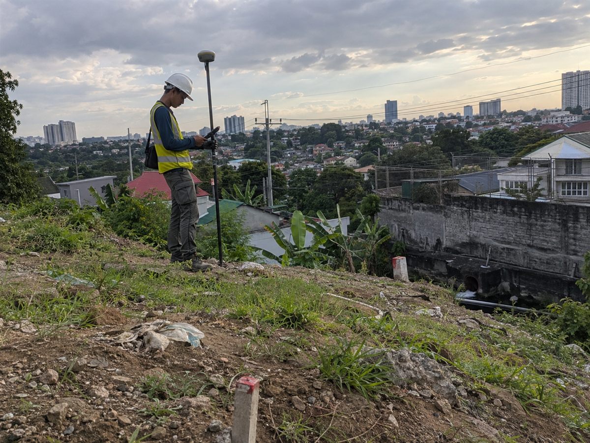 Surveyor in a hard hat using a GNSS rover and handheld controller on a sloped lot with a city skyline in the distance