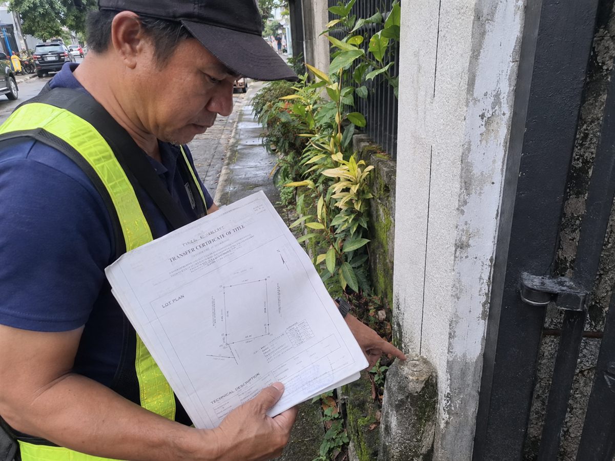 Surveyor in a cap and safety vest holding a Transfer Certificate of Title with a lot plan and pointing at a concrete boundary marker at the base of a wall